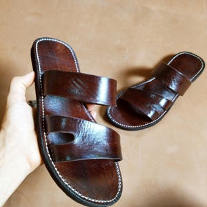 Pu&ograve; includere: Un paio di sandali in pelle marrone con due cinturini e suola cucita. Un sandalo &egrave; tenuto in mano, l'altro poggia su una superficie marrone. Design classico.