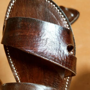 Pu&ograve; includere: Primo piano di un sandalo in pelle marrone con un cinturino sulla parte superiore. Il sandalo ha cuciture bianche sui bordi e una finitura lucida. La pelle sembra invecchiata.