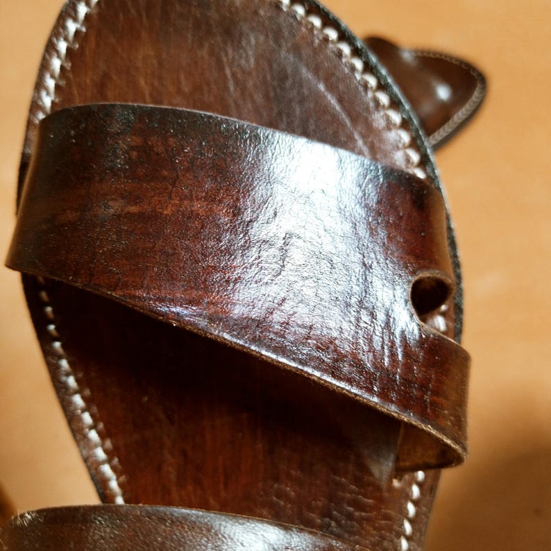 Pu&ograve; includere: Primo piano di un sandalo in pelle marrone con un cinturino sulla parte superiore. Il sandalo ha cuciture bianche sui bordi e una finitura lucida. La pelle sembra invecchiata.