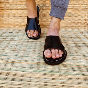 Puede incluir: Sandalias de cuero negro con una correa ancha y costuras blancas. Las sandalias se muestran sobre una estera tejida. Calzado casual, perfecto para el verano.