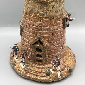 May include: A ceramic lighthouse figurine with a textured brown and tan finish. The lighthouse has a small window and a door with steps leading up to it. There are small figurines of soldiers standing around the base of the lighthouse.