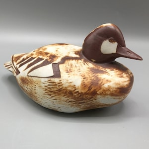 May include: A ceramic duck figurine with brown and white glaze. The duck is in a resting position with its head turned to the side.