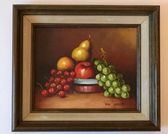 May include: A framed oil painting of a still life featuring a bowl of fruit. The bowl contains a red apple, a yellow pear, and green grapes. There are also red cherries scattered around the bowl. The painting is signed "Van Damm" in the lower right corner.