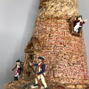 May include: A ceramic model of a lighthouse with three miniature figures of soldiers in red, white, and blue uniforms. The lighthouse is made of brown and red clay with a textured surface.