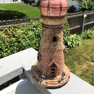 May include: A ceramic lighthouse figurine with a red and white striped roof and a brown base. The lighthouse has a small door and windows, and there are four small figurines of people standing around the base.
