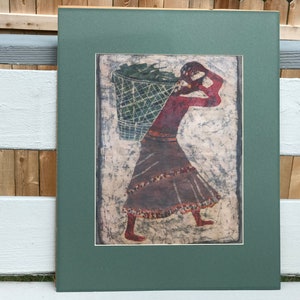 May include: A framed print of a woman carrying a basket of green leaves on her back. The woman is wearing a red top and a brown skirt. The background is a light beige with a textured pattern.