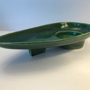May include: A mid-century modern, green ceramic ashtray with a unique, elongated shape. The ashtray features a raised, circular area for holding cigarettes and is supported by three small, rectangular feet. The glossy finish enhances its sleek design.