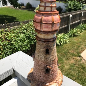 May include: A ceramic lighthouse figurine with a pink and brown glazed finish. The lighthouse has a detailed brick design and two small windows. The top of the lighthouse has a domed roof with a lattice design.