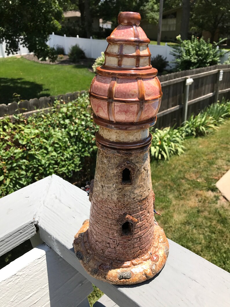 May include: A ceramic lighthouse figurine with a pink and brown glazed finish. The lighthouse has a detailed brick design and two small windows. The top of the lighthouse has a domed roof with a lattice design.