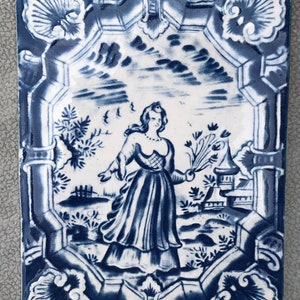 May include: A blue and white ceramic tile with a decorative border and a central image of a woman in a dress holding a bouquet of flowers. The woman is standing in a garden setting with a house in the background.
