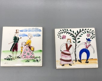 May include: Two square ceramic tiles with hand-painted illustrations. One depicts a couple in period clothing, the other shows two figures dancing under a tree. The tiles have a white background with colorful details.