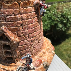 May include: A ceramic tower with a brick facade and two miniature soldiers. One soldier is climbing the tower, while the other is standing at the base.