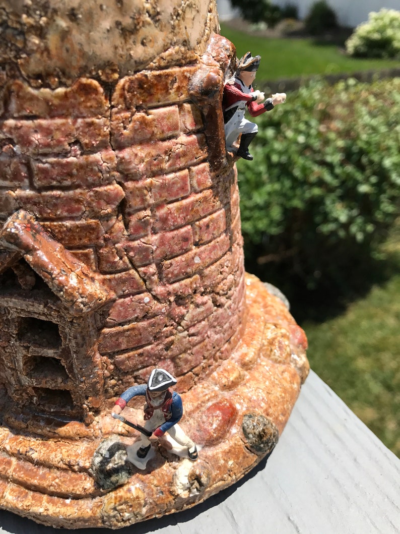 May include: A ceramic tower with a brick facade and two miniature soldiers. One soldier is climbing the tower, while the other is standing at the base.