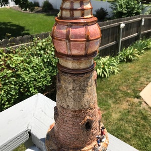 May include: A ceramic lighthouse figurine with a brown and pink glazed finish. The lighthouse has a detailed design with a small window and a tower with a lattice pattern. There are three small figurines at the base of the lighthouse.