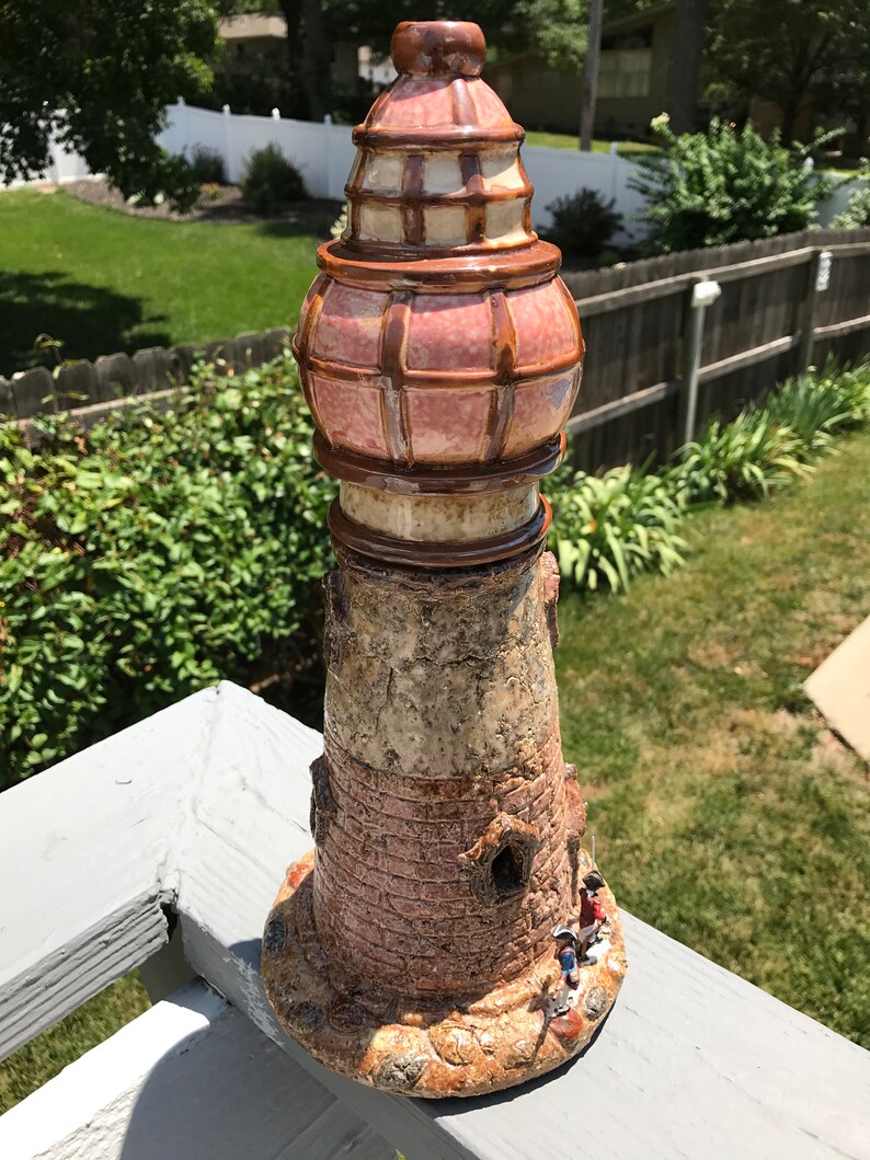 May include: A ceramic lighthouse figurine with a brown and pink glazed finish. The lighthouse has a detailed design with a small window and a tower with a lattice pattern. There are three small figurines at the base of the lighthouse.