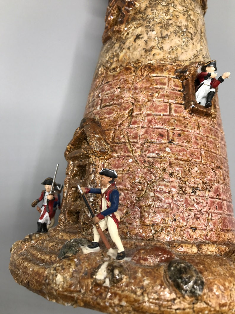 May include: A ceramic model of a lighthouse with three miniature figures of soldiers in red, white, and blue uniforms. The lighthouse is made of brown and red clay with a textured surface.