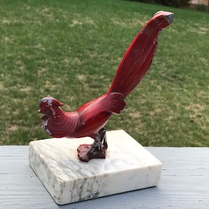 May include: A red metal figurine of a pheasant with a long tail, standing on a white marble base.