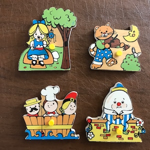 May include: Four wooden puzzle pieces featuring characters from children's stories. The first piece shows a girl in a blue dress sitting on a mushroom under a tree. The second piece shows a cat in a striped shirt playing a violin. The third piece shows three people in a boat. The fourth piece shows a round character in a striped shirt standing on a wall.