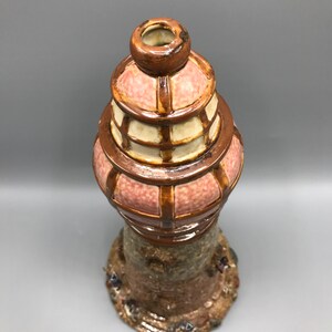 May include: A ceramic vase shaped like a lighthouse with a pink and brown glaze. The vase has a detailed base with small figures.
