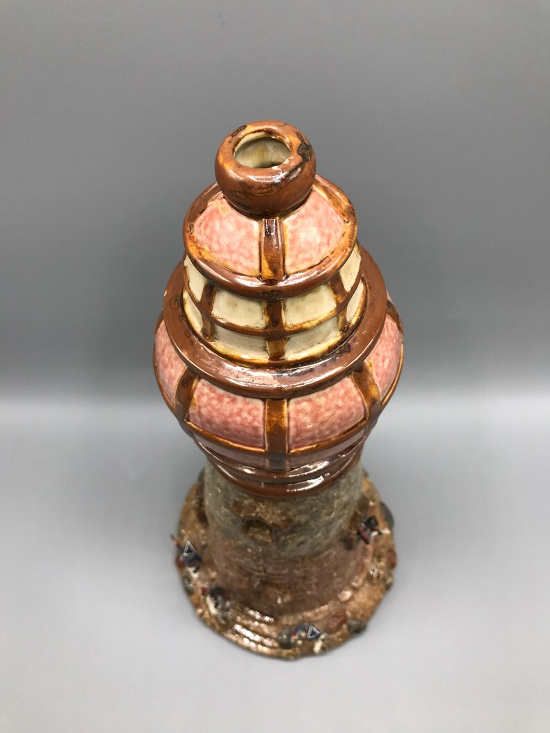 May include: A ceramic vase shaped like a lighthouse with a pink and brown glaze. The vase has a detailed base with small figures.