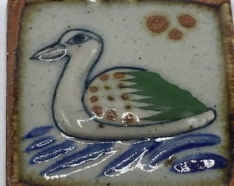 May include: Square ceramic tile featuring a hand-painted duck design. The duck has a white body, green wing, and brown spots. Blue lines represent water. The tile has a brown border and a speckled background.