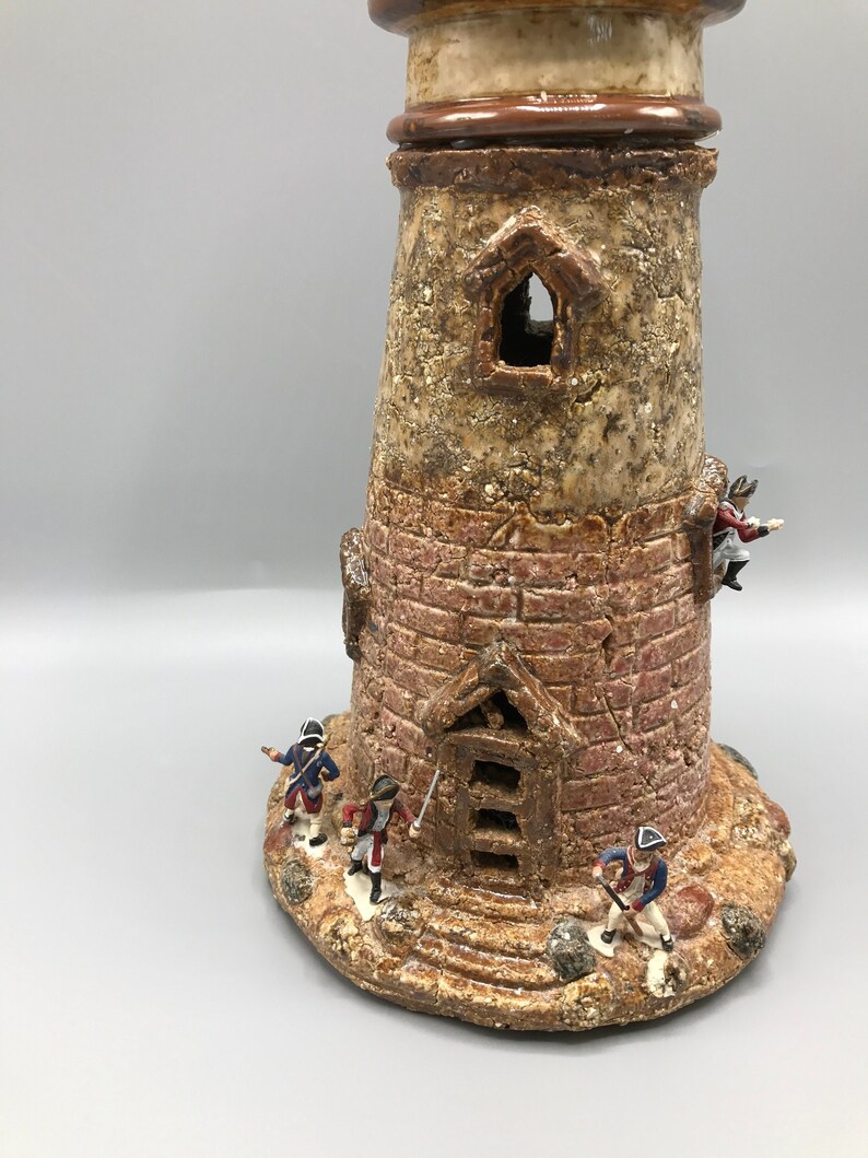 May include: A ceramic lighthouse figurine with a textured brown and tan finish. The lighthouse has a small window and a door with steps leading up to it. There are small figurines of soldiers standing around the base of the lighthouse.