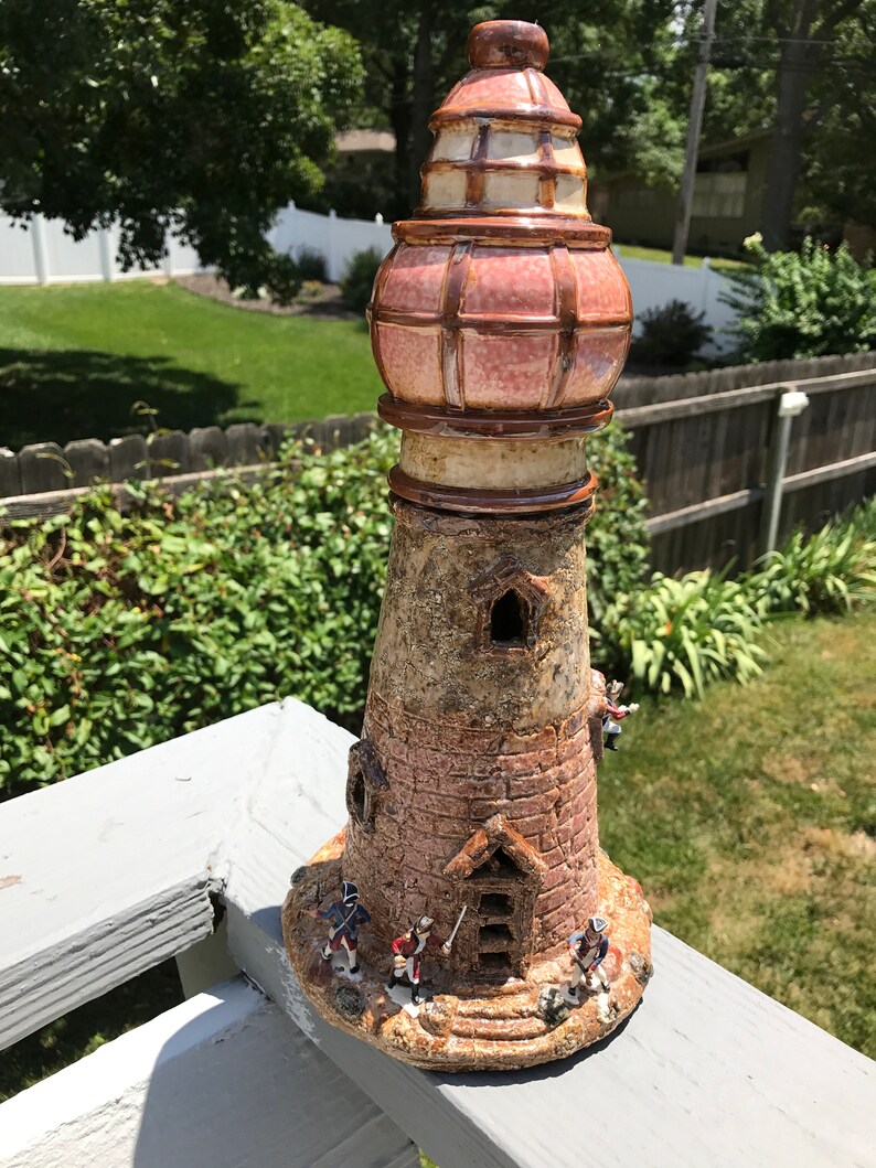 May include: A ceramic lighthouse figurine with a red and white striped roof and a brown base. The lighthouse has a small door and windows, and there are four small figurines of people standing around the base.
