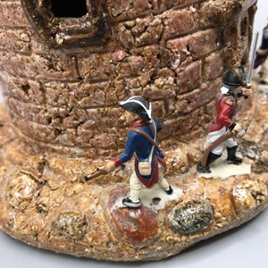 May include: A ceramic miniature castle with three toy soldiers standing on the base. The castle is made of brown and tan clay and has a rough texture. The soldiers are wearing blue and red uniforms and are holding muskets.