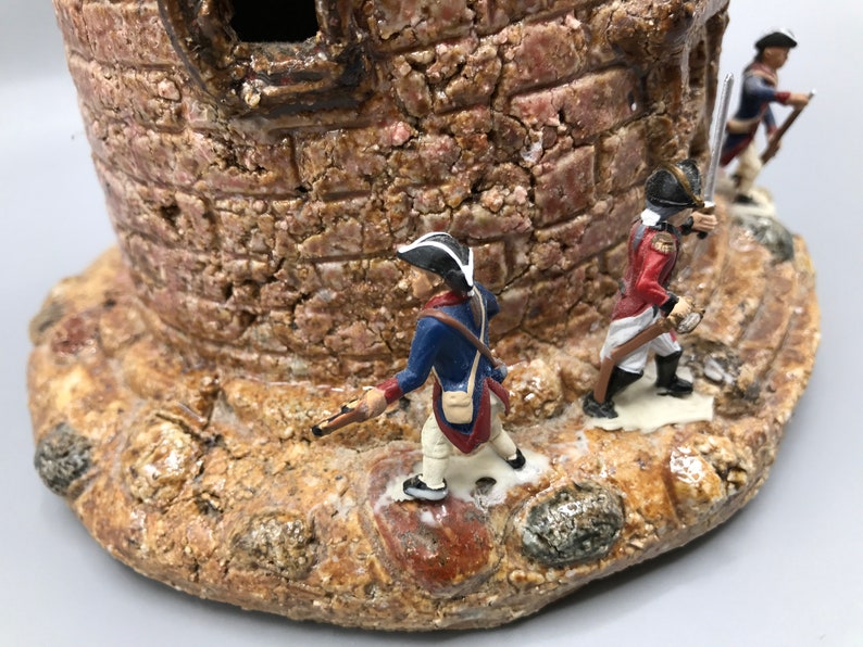 May include: A ceramic miniature castle with three toy soldiers standing on the base. The castle is made of brown and tan clay and has a rough texture. The soldiers are wearing blue and red uniforms and are holding muskets.
