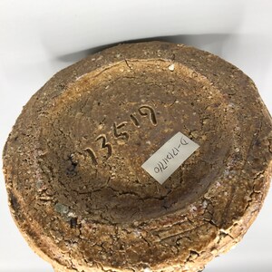 May include: The bottom of a brown ceramic vase with a rough texture. The vase has a number "13519" and a label that says "D-121410" written on it.