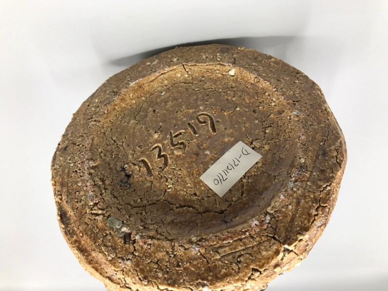 May include: The bottom of a brown ceramic vase with a rough texture. The vase has a number "13519" and a label that says "D-121410" written on it.
