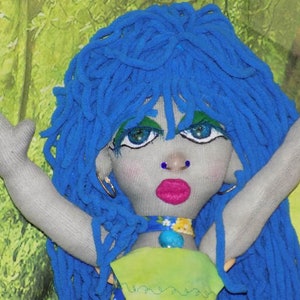 May include: A handmade doll with blue yarn hair, gray fabric arms, and a painted face. The doll is wearing a green and blue fabric dress with a turquoise necklace.
