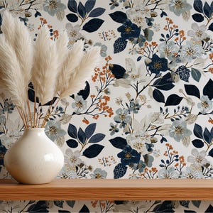 May include: A cream-colored vase holds tall, feathery pampas grass. The background is a floral wallpaper with blue, gray, and rust-colored flowers and leaves. The vase sits on a wooden shelf.