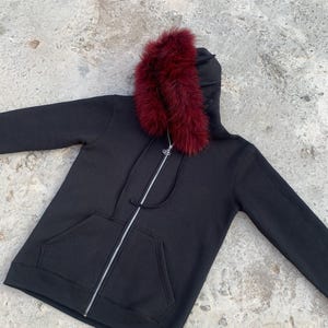 Könnte beinhalten: Ein schwarzer Kapuzenpullover mit einem bordeauxfarbenen Kunstfellbesatz an der Kapuze. Der Pullover hat einen durchgehenden silbernen Reißverschluss und eine Kängurutasche auf der Vorderseite. Das Kleidungsstück ist aus weichem, bequemem Material.