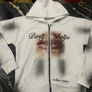 May include: White zip-up hoodie with a hood and front pockets. The hoodie features the words "Dirty Boys" above a graphic of lips. The text "**Rush Archives" is printed below the graphic. The sleeves have a dark grey design.