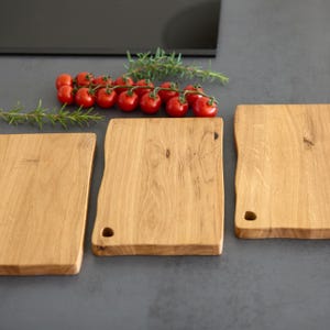May include: Three wooden cutting boards with a natural wood grain finish are arranged on a gray surface. A sprig of rosemary and a cluster of red cherry tomatoes are placed nearby. Each board has a unique, organic shape.