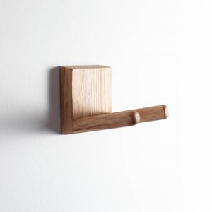 Puede incluir: Un gancho de pared de madera con una base cuadrada y un brazo horizontal redondeado. El gancho est&aacute; hecho de madera de color claro y est&aacute; montado en una pared blanca. El dise&ntilde;o es minimalista y moderno, adecuado para colgar objetos.