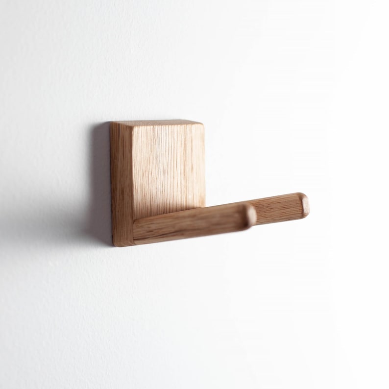 Puede incluir: Un gancho de pared de madera con una base cuadrada y un brazo horizontal redondeado. El gancho est&aacute; hecho de madera de color claro y est&aacute; montado en una pared blanca. El dise&ntilde;o es minimalista y moderno, adecuado para colgar objetos.