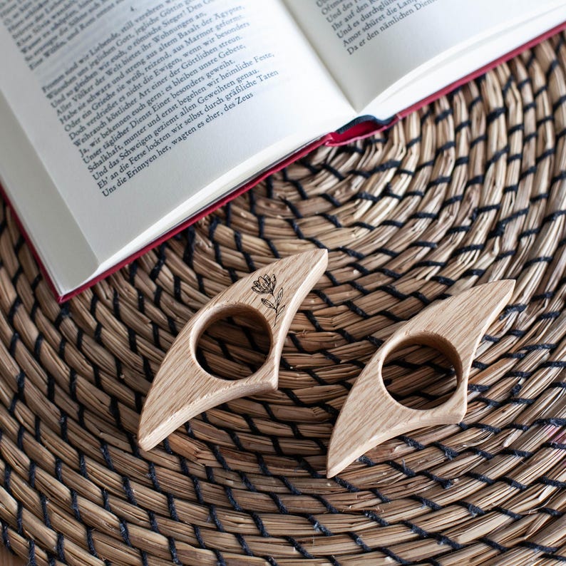 K&ouml;nnte beinhalten: Zwei h&ouml;lzerne Daumen-Buchst&uuml;tzen mit floralem Design, die auf einer gewebten Matte neben einem offenen Buch liegen. Die Buchst&uuml;tzen sind aus hellem Holz gefertigt und haben eine kreisf&ouml;rmige &Ouml;ffnung f&uuml;r den Daumen.