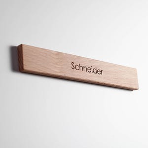 Könnte beinhalten: Ein hellfarbenes Holzschild mit dem eingravierten Namen "Schneider" in einer einfachen Schriftart. Das Schild ist rechteckig mit abgeschrägter Kante und an einer weißen Wand befestigt. Die Holzmaserung ist sichtbar.