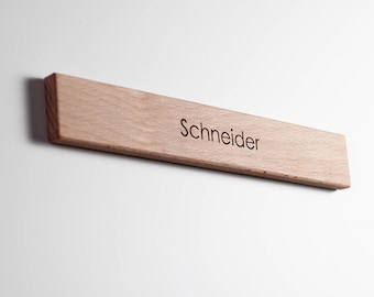 Könnte beinhalten: Ein hellfarbenes Holzschild mit dem eingravierten Namen "Schneider" in einer einfachen Schriftart. Das Schild ist rechteckig mit abgeschrägter Kante und an einer weißen Wand befestigt. Die Holzmaserung ist sichtbar.