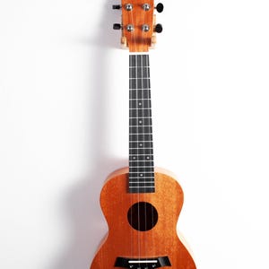 Puede incluir: Un ukelele de madera con un acabado natural. El instrumento tiene un cuerpo redondo, una boca y un diapas&oacute;n con trastes negros. El ukelele est&aacute; colgado sobre un fondo blanco.