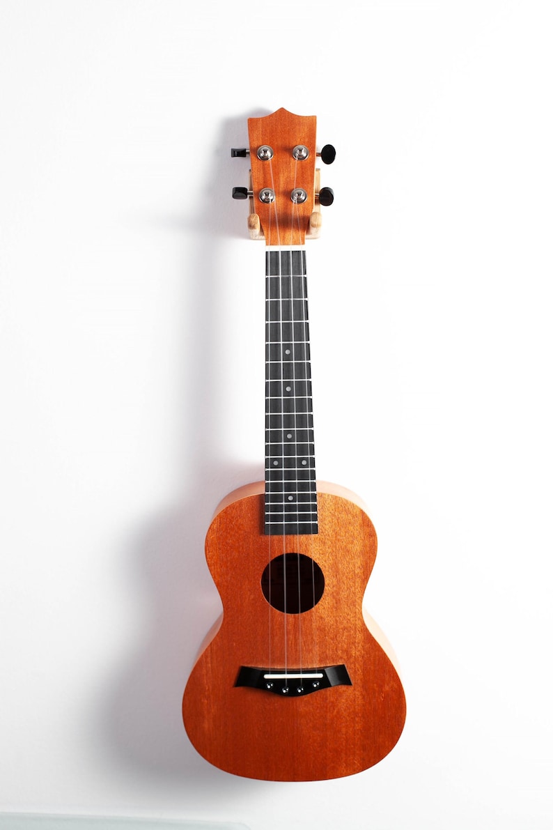 Puede incluir: Un ukelele de madera con un acabado natural. El instrumento tiene un cuerpo redondo, una boca y un diapas&oacute;n con trastes negros. El ukelele est&aacute; colgado sobre un fondo blanco.