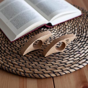 K&ouml;nnte beinhalten: Zwei h&ouml;lzerne Daumen-Buchst&uuml;tzen mit floralem Design, die auf einer gewebten Matte neben einem offenen Buch liegen. Die Buchst&uuml;tzen sind hellbraun und haben eine kreisf&ouml;rmige &Ouml;ffnung f&uuml;r den Daumen.