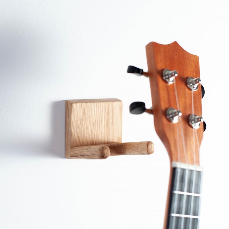 Puede incluir: Un soporte de pared de madera para ukelele con acabado natural. El soporte es cuadrado con un brazo curvado para sujetar el instrumento. Se ve parte de un ukelele, mostrando el clavijero y las cuerdas.
