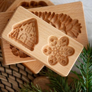 Könnte beinhalten: Holz-Keksformen mit geschnitzten Designs, darunter ein Haus, ein Schneeflocken- und ein Blumenmuster. Die Formen sind gestapelt und zeigen die detaillierte Handwerkskunst und die natürliche Holzmaserung. Ideal zum Backen.