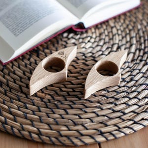 K&ouml;nnte beinhalten: Zwei h&ouml;lzerne Daumen-Buchst&uuml;tzen mit Rosenmuster, die auf einer gewebten Matte neben einem offenen Buch liegen. Die Buchst&uuml;tzen sind hellbraun mit einem kreisf&ouml;rmigen Loch f&uuml;r den Daumen und einem geschwungenen Design.
