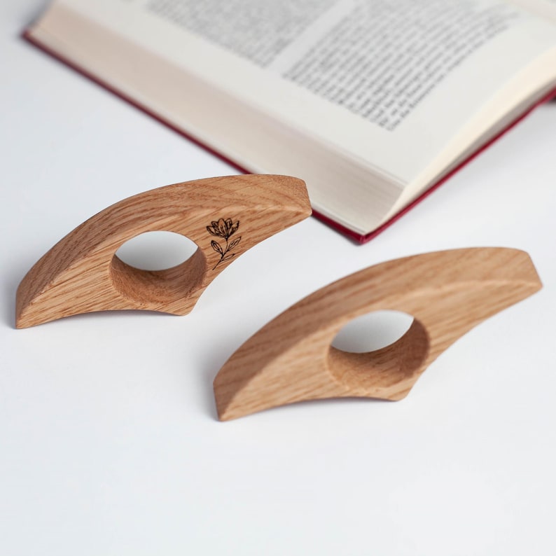 K&ouml;nnte beinhalten: Zwei h&ouml;lzerne Buchst&uuml;tzen mit floralem Design. Die hellbraunen Halterungen haben eine kreisf&ouml;rmige &Ouml;ffnung f&uuml;r den Finger. Im Hintergrund ist ein Buch mit roten Kanten zu sehen.