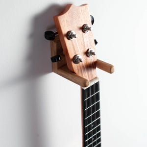 Puede incluir: Un ukelele de madera marr&oacute;n claro montado en una pared. El instrumento tiene un diapas&oacute;n negro y clavijas de afinaci&oacute;n plateadas. El ukelele est&aacute; sujeto por un soporte de madera hecho a medida.