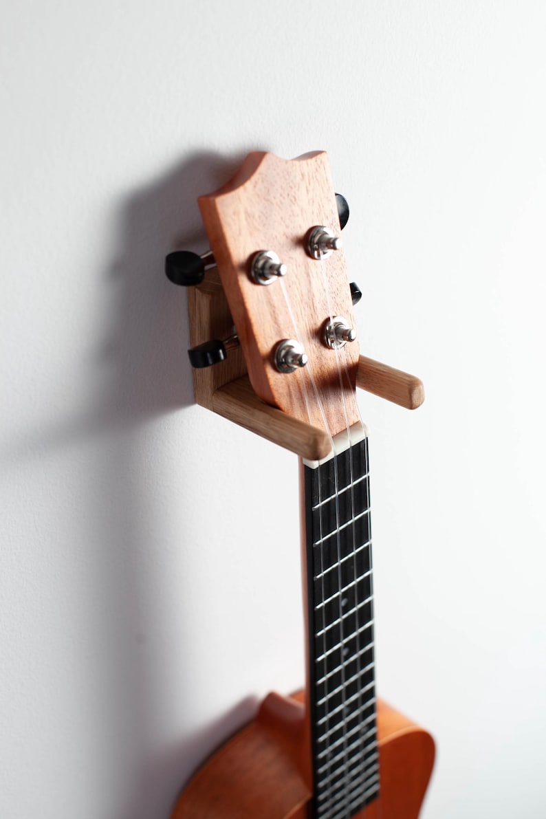Puede incluir: Un ukelele de madera marr&oacute;n claro montado en una pared. El instrumento tiene un diapas&oacute;n negro y clavijas de afinaci&oacute;n plateadas. El ukelele est&aacute; sujeto por un soporte de madera hecho a medida.